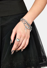 A close-up of a person's hand wearing multiple silver-toned jewelry pieces, including a heart-shaped ring and layered chain bracelets, styled with a black dress against a light gray background.