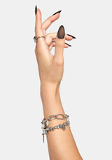 A close-up of a person's hand and wrist wearing multiple silver-toned bracelets, including a chunky chain bracelet and a another bracelet with a dagger charm, against a light gray background.