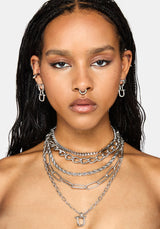 A close-up of a model wearing a layered stack of silver-tone chain necklaces, including chunky cable, paperclip, and delicate link styles, along with matching silver hoop earrings.