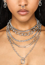 A close-up of a model wearing a layered silver chain necklace set with a U-shaped pendant, along with matching silver hoop earrings and a nose ring.