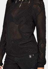 A close-up of a person wearing a black sheer mesh long-sleeve top with large tonal embroidered or appliqué details on the front, paired with black pants.