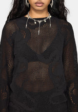 A close-up of a model wearing a black sheer crochet knit top with tonal floral lace applique overlays, styled with silver statement jewelry against a light gray background.