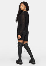 A woman modeling a black long-sleeve sheer mesh top with black shorts and black over-the-knee boots, shown from a side-back angle against a light gray studio background.