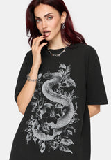 A woman wearing an oversized black t-shirt with a detailed white graphic of a snake wrapped through floral elements, set against a plain light gray background.