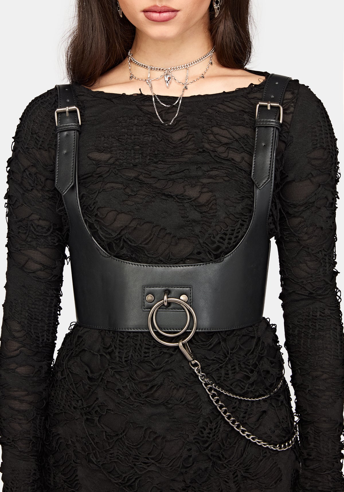 A close-up of a model wearing a black lace long-sleeve dress with a black faux-leather corset-style waist harness featuring silver-tone metal buckles, a large front ring, and a hanging chain.
