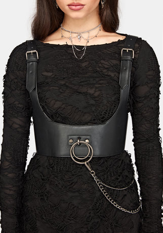 A close-up of a model wearing a black lace long-sleeve dress with a black faux-leather corset-style waist harness featuring silver-tone metal buckles, a large front ring, and a hanging chain.
