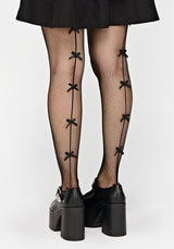 Black fishnet tights with decorative bow details down the front, worn with black platform Mary Jane shoes and a black skirt against a light gray background.