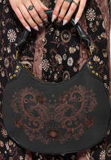 Black handbag with intricate brown embroidery held by a person against a patterned fabric background