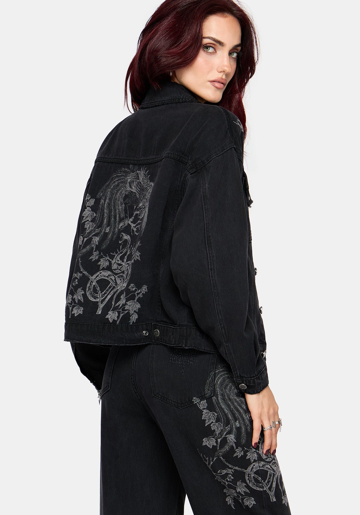 A woman wearing a black denim jacket with faded gray graphic prints on the back and upper thigh, paired with black jeans, photographed against a light gray studio background.
