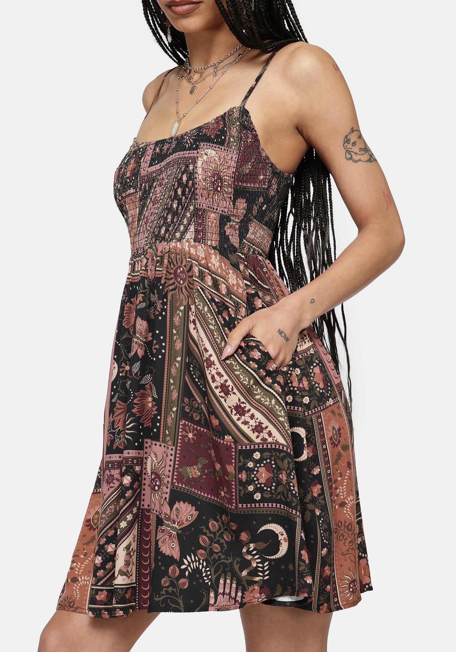 A woman wearing a sleeveless spaghetti-strap mini dress with a bohemian patchwork print in dusty pink, rust, brown, and black, shown against a light gray studio background.