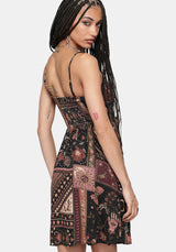 A woman modeling a sleeveless spaghetti-strap mini dress with a bohemian patchwork print in black, rust red, and beige, shown from a three-quarters back view against a light gray background.