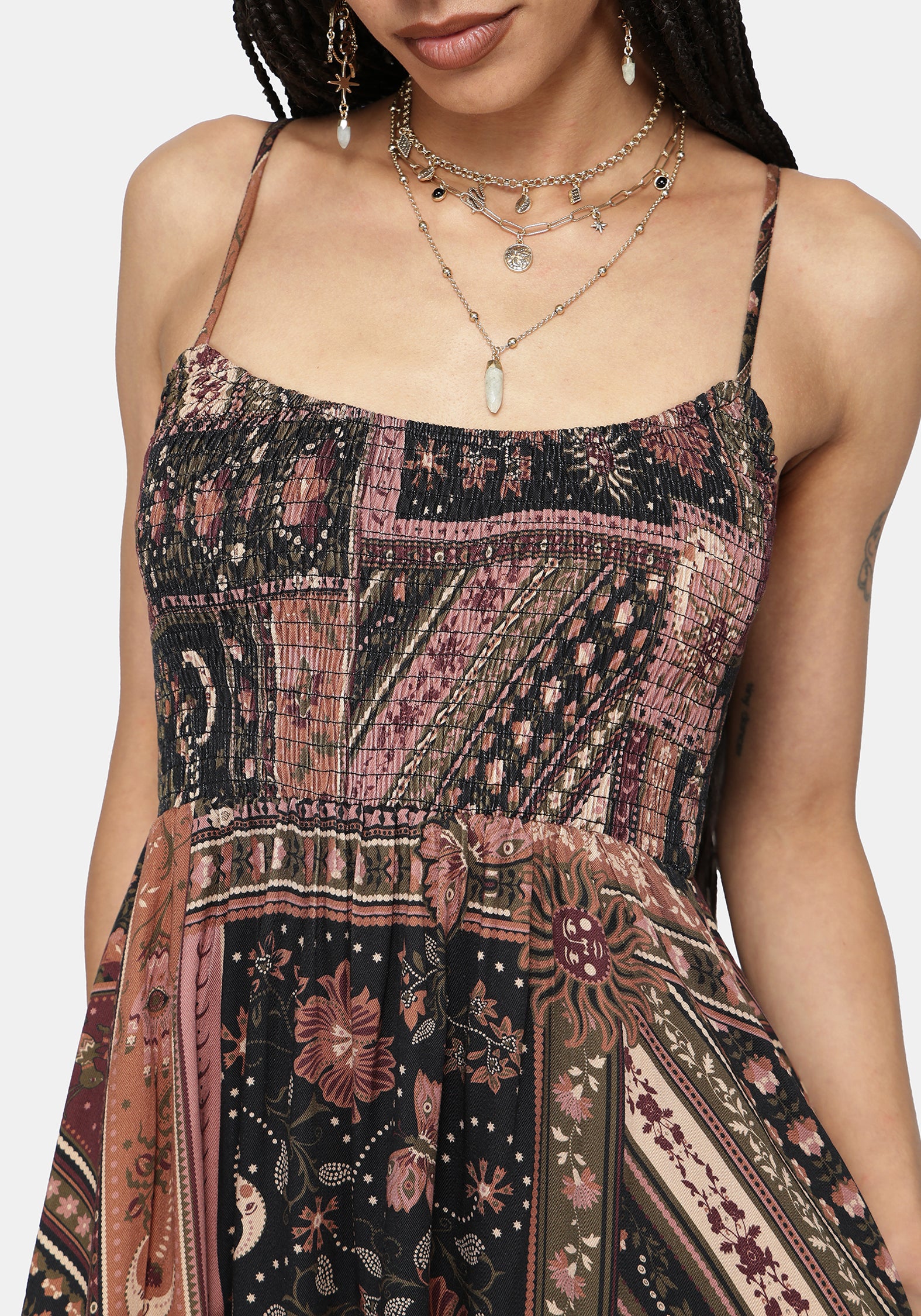 A close-up of a woman wearing a sleeveless patchwork-print dress with thin straps and a tie waist, shown against a plain light background. The dress has a multicolor print with pink, black, and brown tones.