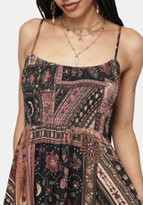 A close-up of a woman wearing a sleeveless patchwork-print dress with thin straps and a tie waist, shown against a plain light background. The dress has a multicolor print with pink, black, and brown tones.
