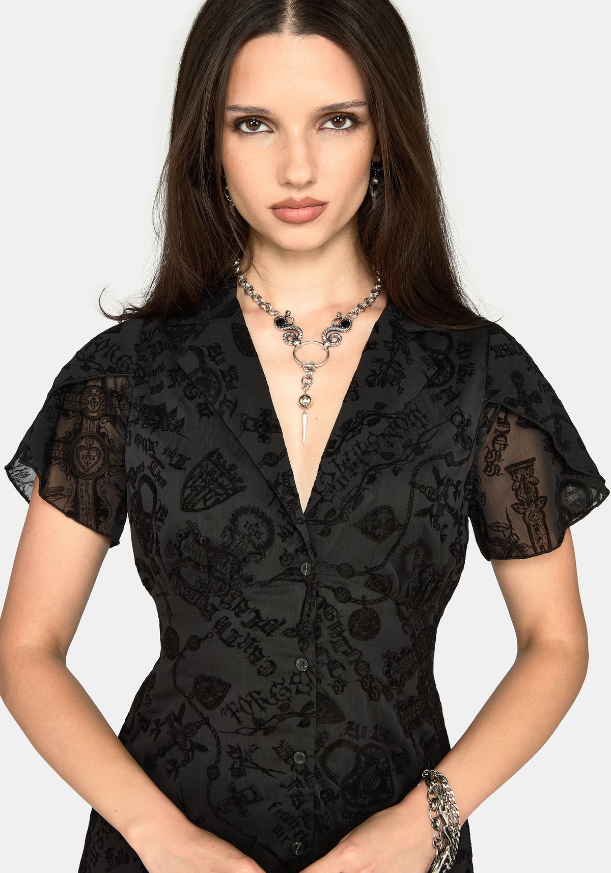 A woman wearing a black patterned button-front dress with sheer flutter sleeves, photographed against a light gray studio background.