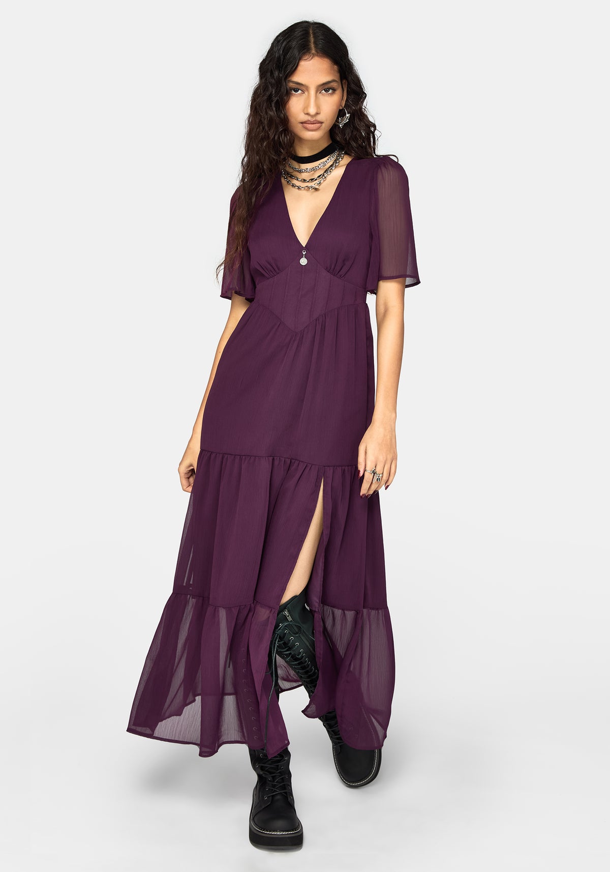 A woman modeling a long, deep purple maxi dress with sheer flutter sleeves, a V-neckline, and a tiered skirt with a high side slit, styled with black boots and layered necklaces against a light gray studio background.