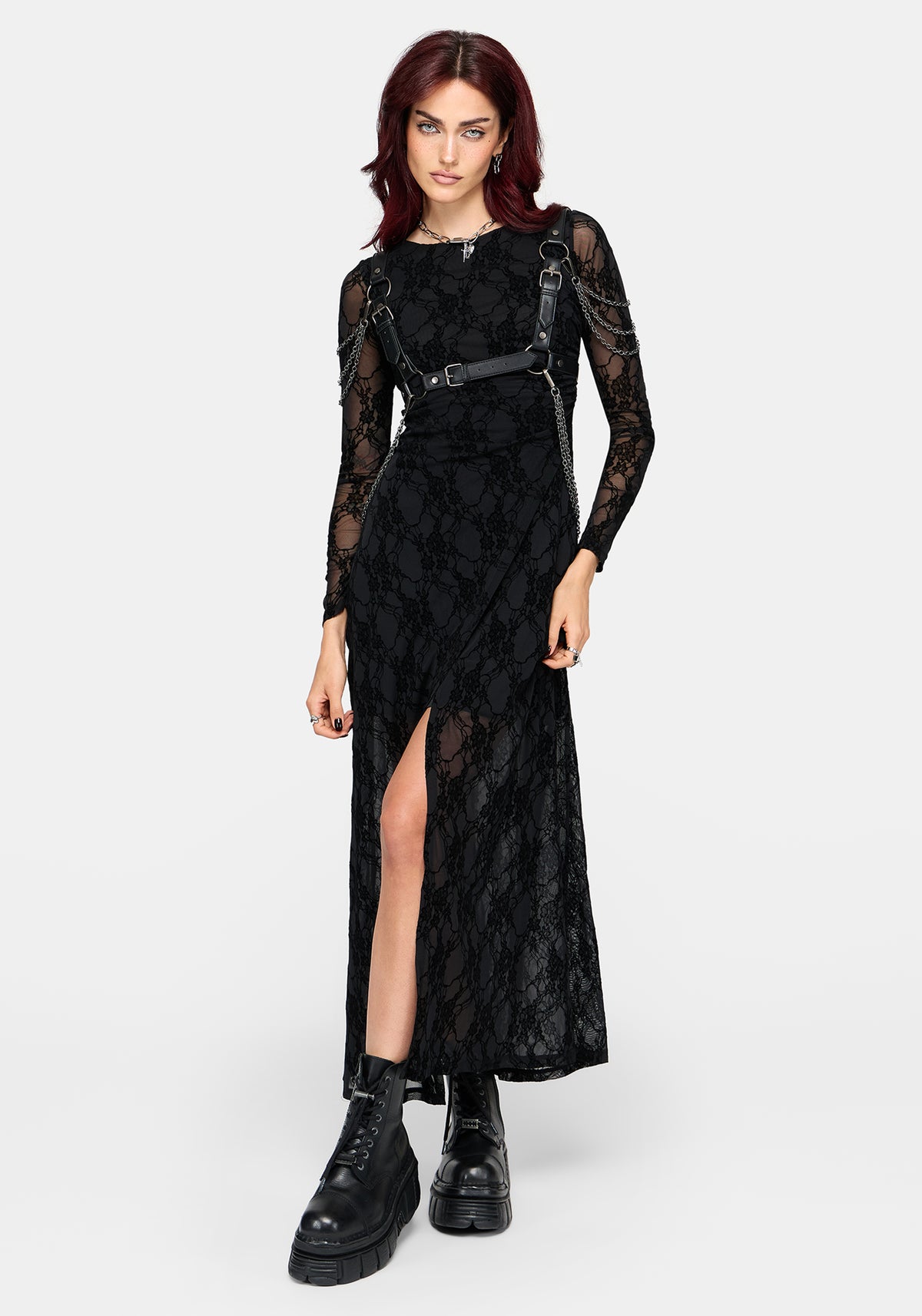 A model wearing a black lace long-sleeve maxi dress with a thigh-high slit, a matching black faux-leather belt with metal buckles across the waist, and black combat boots, set against a plain light gray studio background.