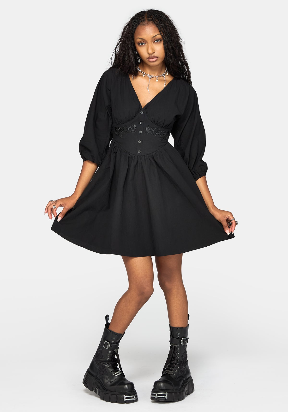 A woman modeling a black short dress with three-quarter sleeves, a V-neckline, and a buttoned waistband, paired with black combat boots against a light gray studio background.