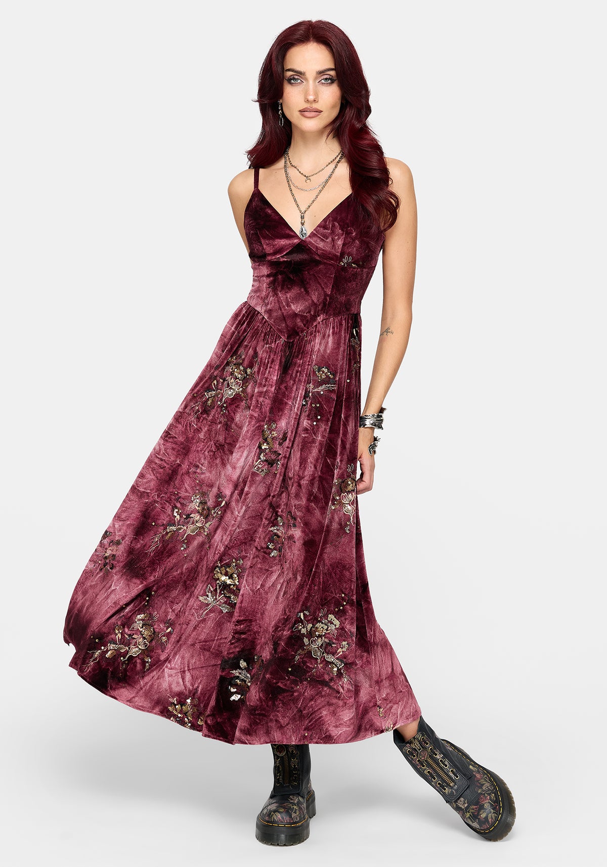 A woman modeling a sleeveless, floor-length dusty rose velvet dress with a floral pattern and tie-dye texture, paired with black boots, against a light gray studio background.