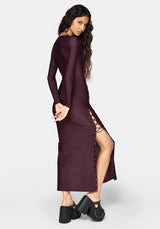 A woman modeling a long-sleeve, deep plum purple midaxi dress with a high side slit and lace-up detail, paired with black platform clogs, against a light gray studio background.
