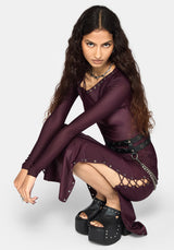 A model crouching in a studio setting, wearing a dark plum long-sleeve dress with silver studs, a matching skirt, a black studded belt, and black platform sandals.