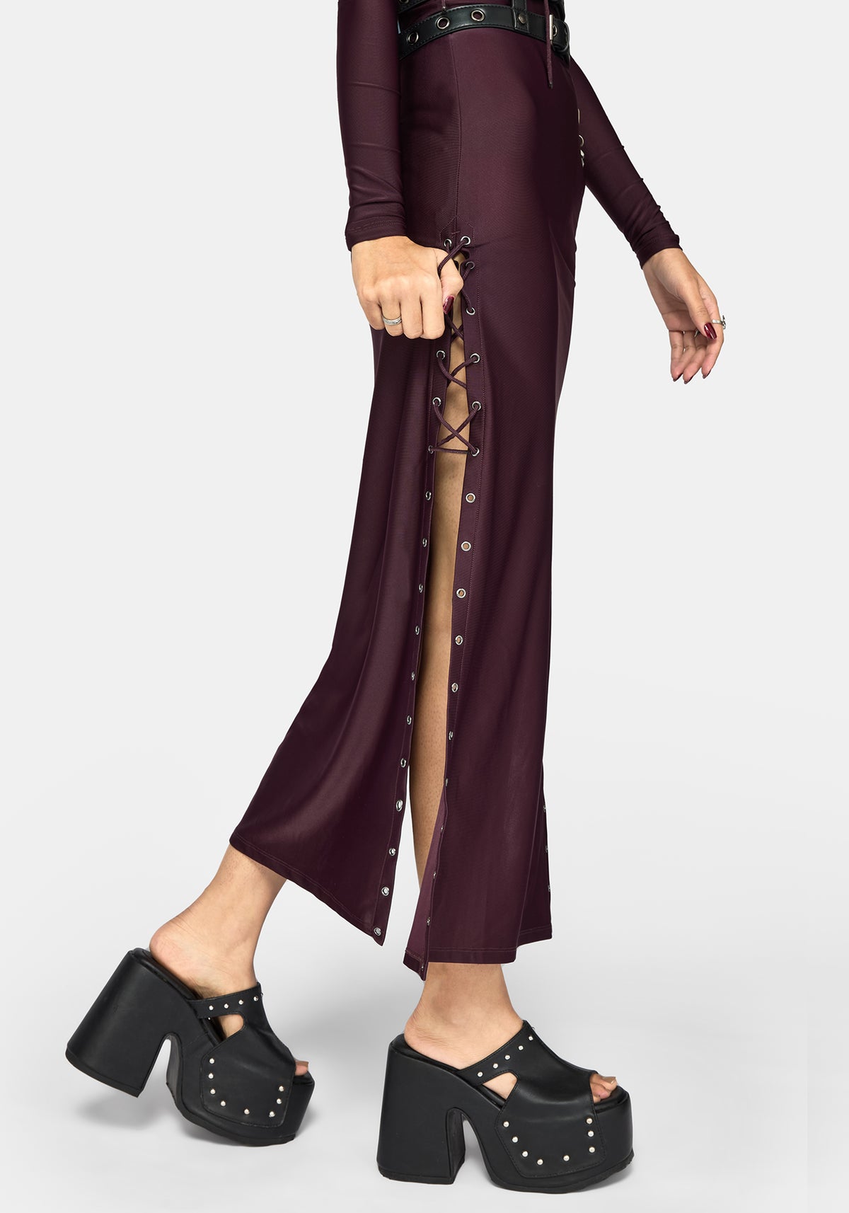 A model wearing a long-sleeved, deep plum purple maxi dress with a high side slit, lace-up front detail, and silver studs, paired with chunky black platform sandals, against a light gray background.