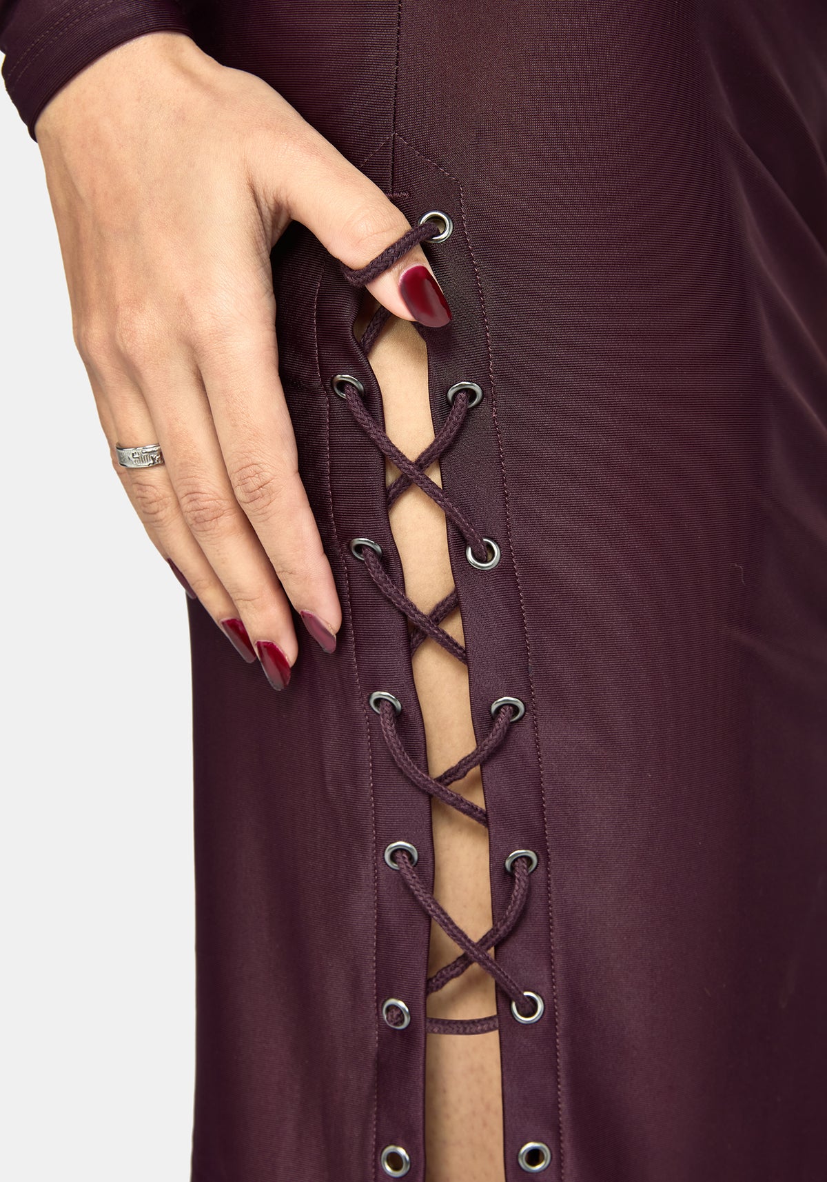 Close-up of a person wearing a dark plum lace-up dress, showing a hand with dark red nail polish adjusting the side laces with silver metal eyelets.