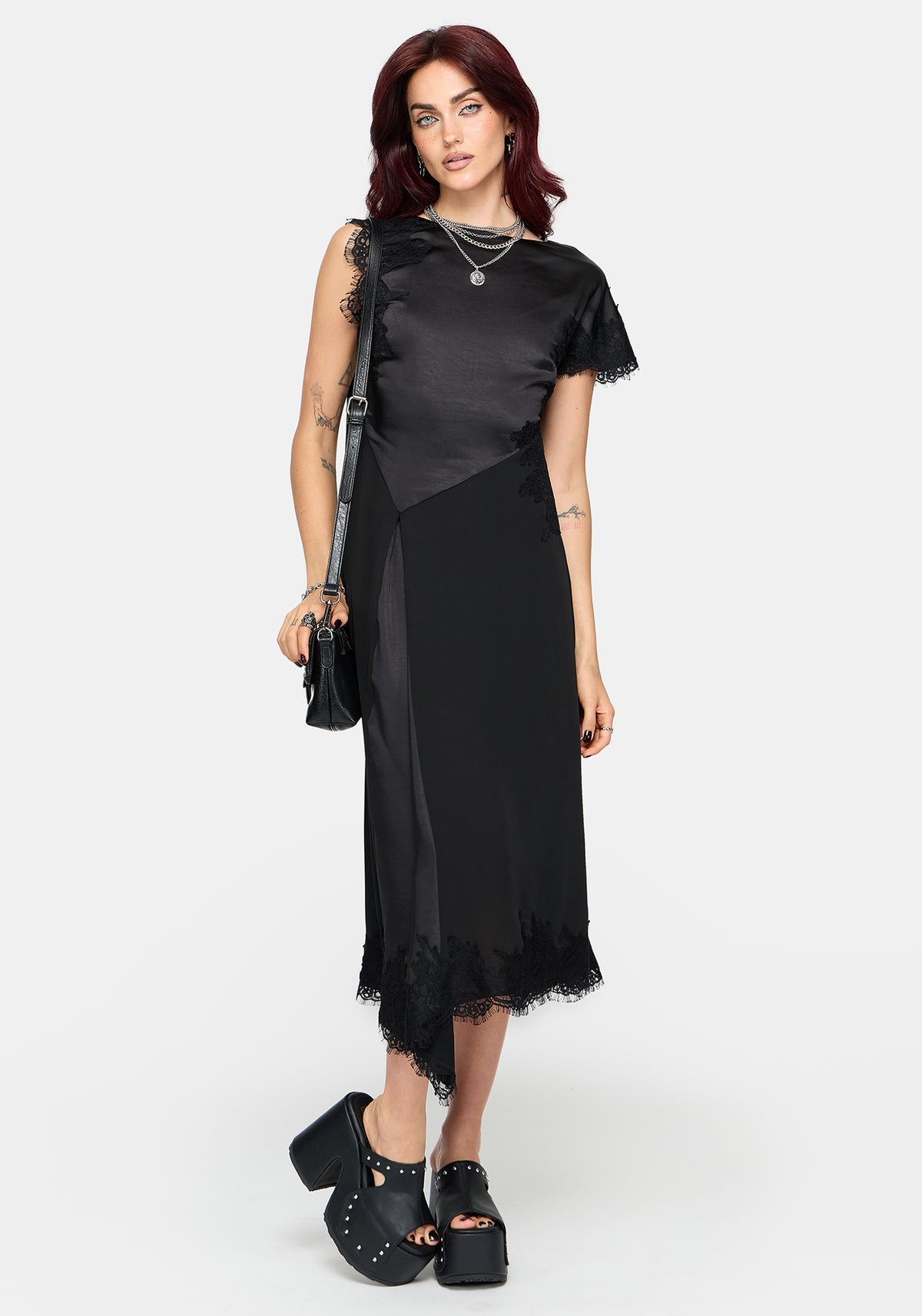A woman modeling a black midi dress with sheer mesh side panels and tattered lace trim, styled with layered silver necklaces, a black shoulder bag, and black platform sandals against a light gray studio background.