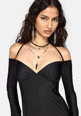 A woman modeling a black off-the-shoulder long-sleeve dress with layered silver and black necklaces, against a light gray background.