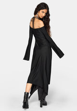 A woman modeling a black off-the-shoulder long-sleeve midi dress with an asymmetrical hem and black ankle boots, photographed against a light gray studio background.