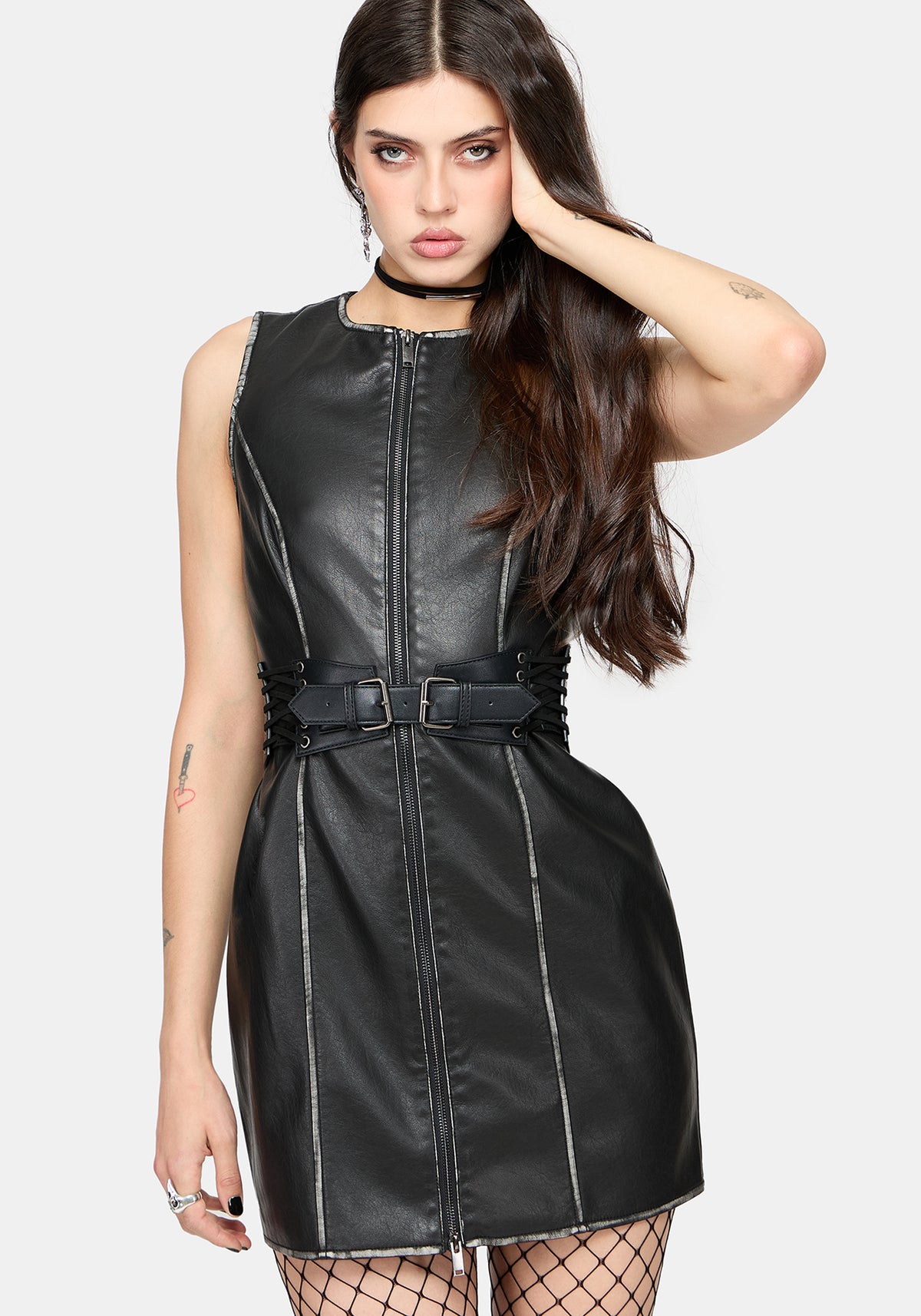 A woman modeling a black faux-leather sleeveless mini dress with a front zipper, side lacing, and a matching belt with a buckle, against a light gray studio background.