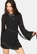 A woman modeling a black long-sleeve mini dress with sheer bell sleeves, a studded waist tie belt, and studded trim along the hem, against a light gray studio background.