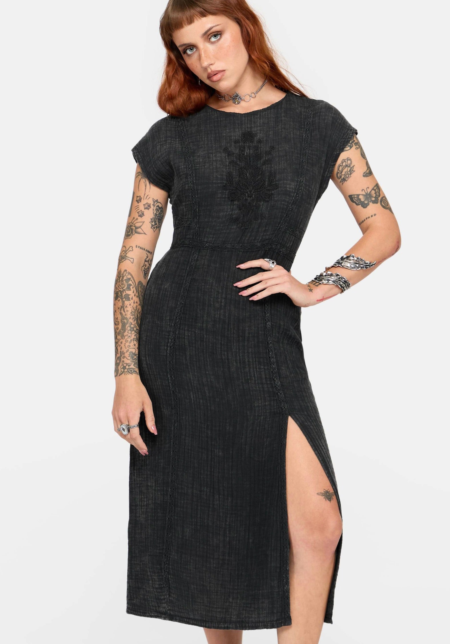 A woman modeling a black textured midi dress with short cap sleeves, a round neckline, and a thigh-high side slit, photographed against a plain light gray studio background.