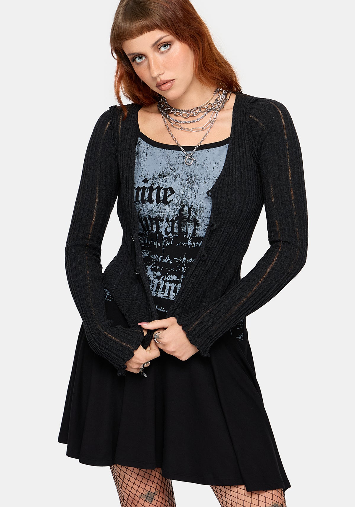 A model wearing a black long-sleeve ribbed cardigan over a black and gray graphic print mini dress and black, styled with layered silver necklaces and fishnet tights against a light gray background.