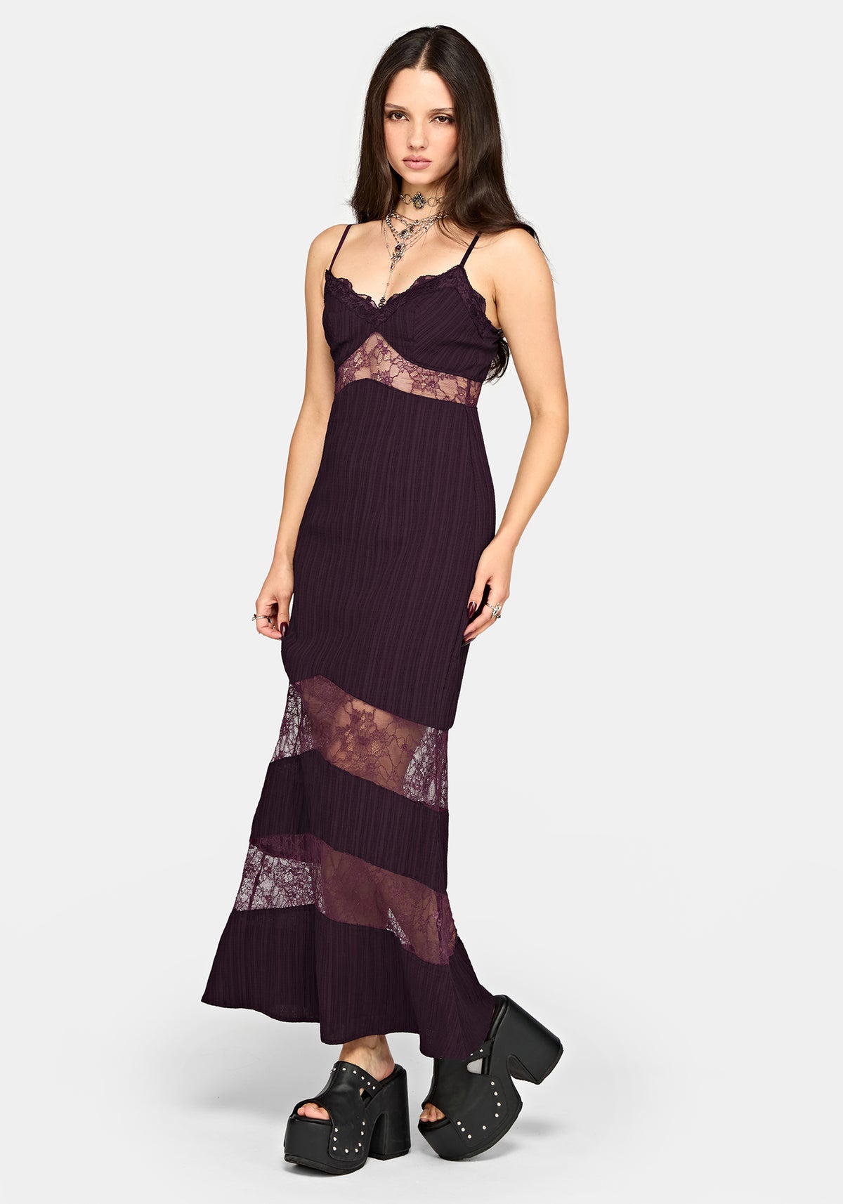 A woman modeling a dark plum purple maxi dress with pleated fabric, lace inserts, and black platform shoes against a light gray studio background.