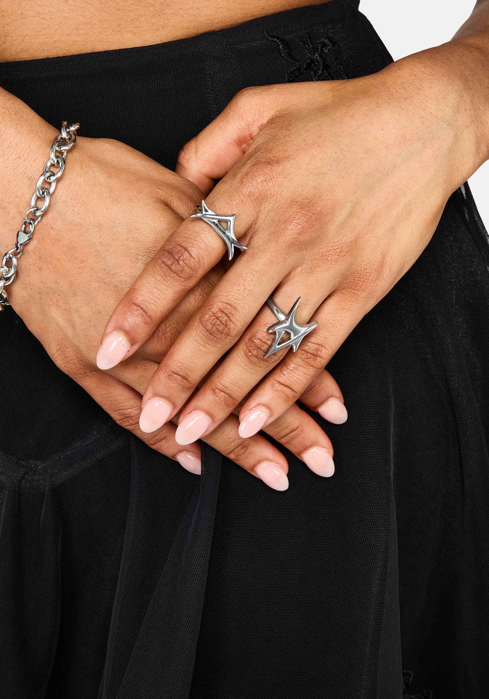 A close-up of a person's hands wearing two silver rings with spike designs, one on the ring finger and one on the middle finger, along with a silver chain bracelet on the wrist, set against a black skirt.