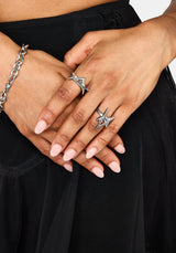 A close-up of a person's hands wearing two silver rings with spike designs, one on the ring finger and one on the middle finger, along with a silver chain bracelet on the wrist, set against a black skirt.