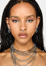 A close-up portrait of a model wearing multiple layered silver chain necklaces, matching silver hoop earrings, and a silver nose ring against a light gray background.