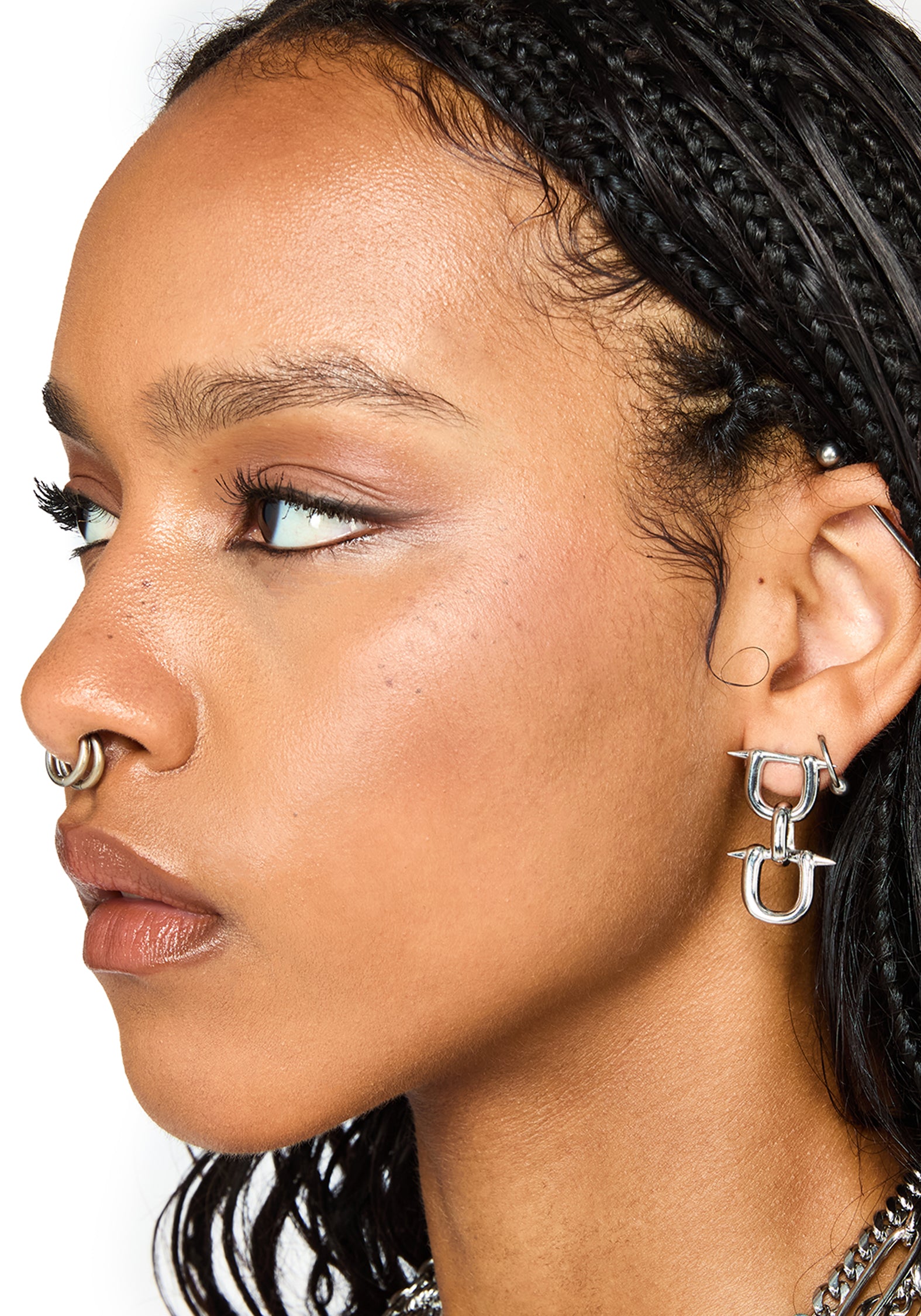 Close-up side profile of a woman wearing a silver double-U hoop earring on her ear, a nose ring, and layered necklaces against a white background.