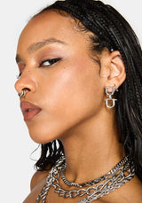 A close-up side profile of a model wearing a silver geometric hoop earring, a nose ring, and layered silver chain necklaces against a light gray background.