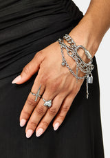 A close-up of a person's hand and wrist wearing multiple silver-toned chain bracelets, including a chunky chain bracelet with a padlock charm and a ring with a sword-shaped setting, against a black dress and light gray background.