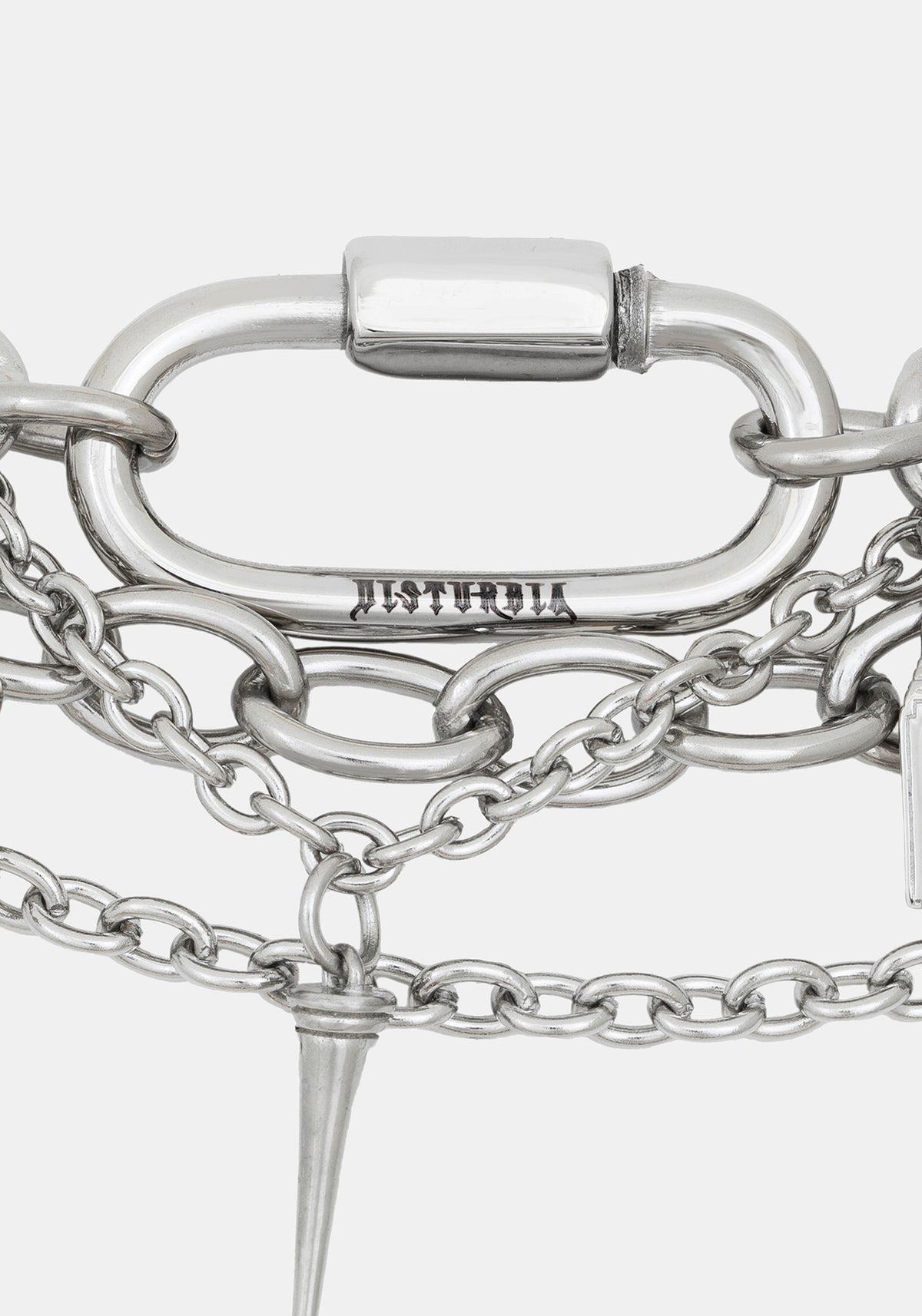 Close-up of a silver-toned chain bracelet featuring a large oval carabiner clasp engraved with 'DISTURBIA', layered chain links, and a pointed spike charm, on a light gray background.