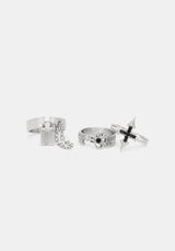 Three silver-toned gothic-style rings displayed on a light gray background, including a cross ring with black accents, a ring with a hanging chain and padlock charm, and a ring with a skull detail.