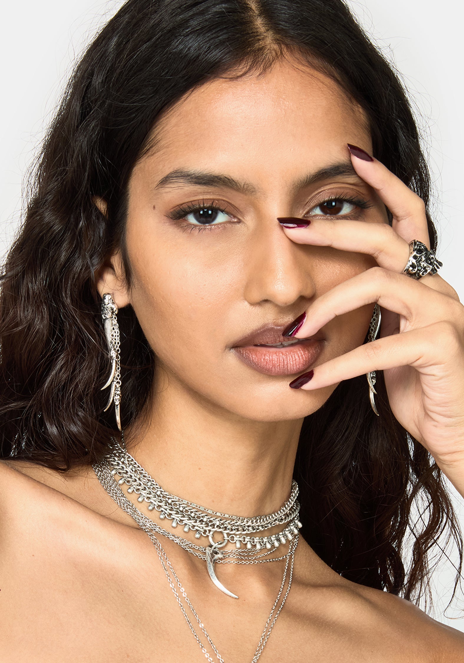 A close-up of a woman wearing layered silver jewelry, including a multi-strand choker necklace, a long feather earring, and a ring, against a plain white background.