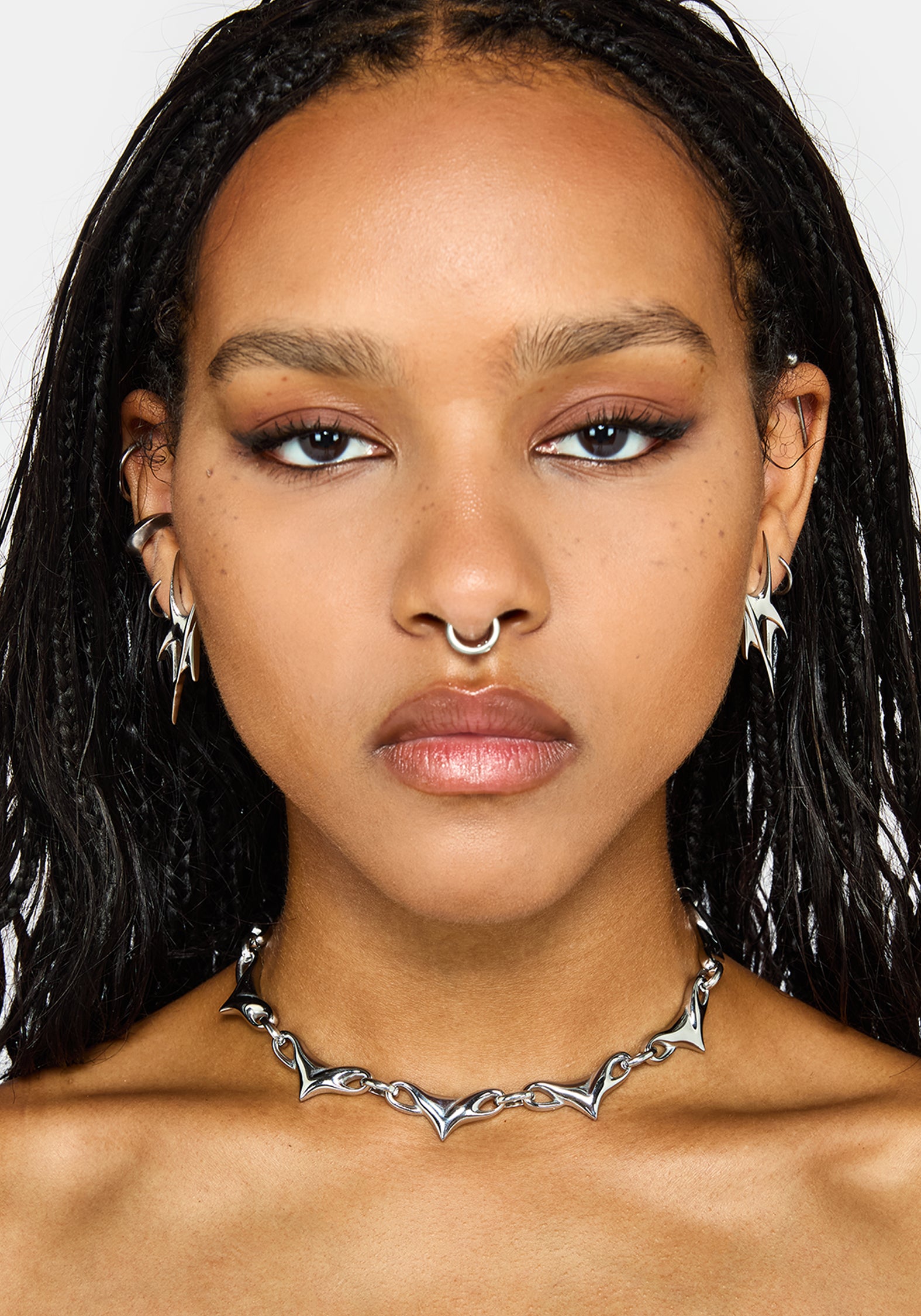 A close-up portrait of a model wearing a silver spiked choker necklace, matching silver hoop nose ring, and spike stud earrings against a light gray background.