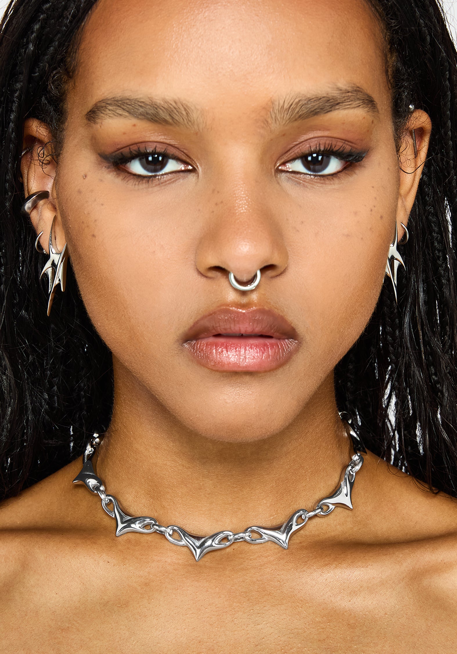 A close-up portrait of a model wearing a silver choker necklace with a spike, jagged, bat-wing-like design. The model also has other silver piercings and hoop earrings.