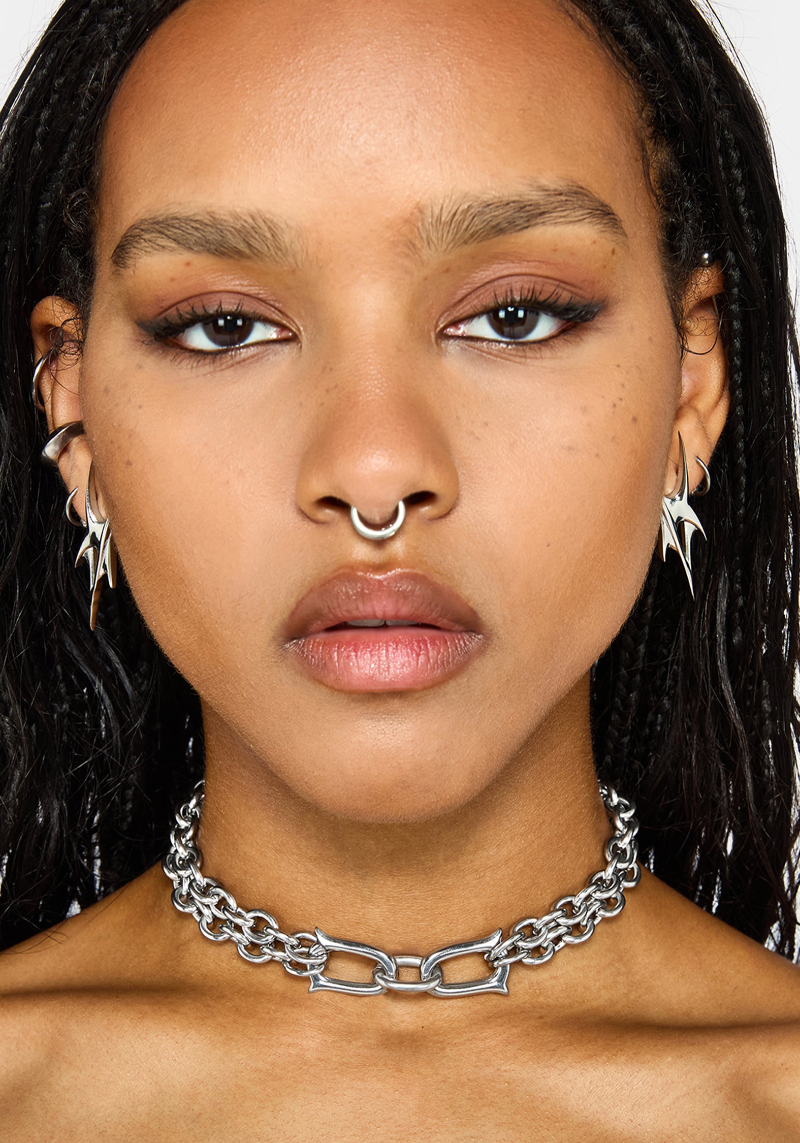 A close-up of a model wearing a chunky silver chain choker, along with matching silver hoop earrings and a nose ring, against a plain white background.