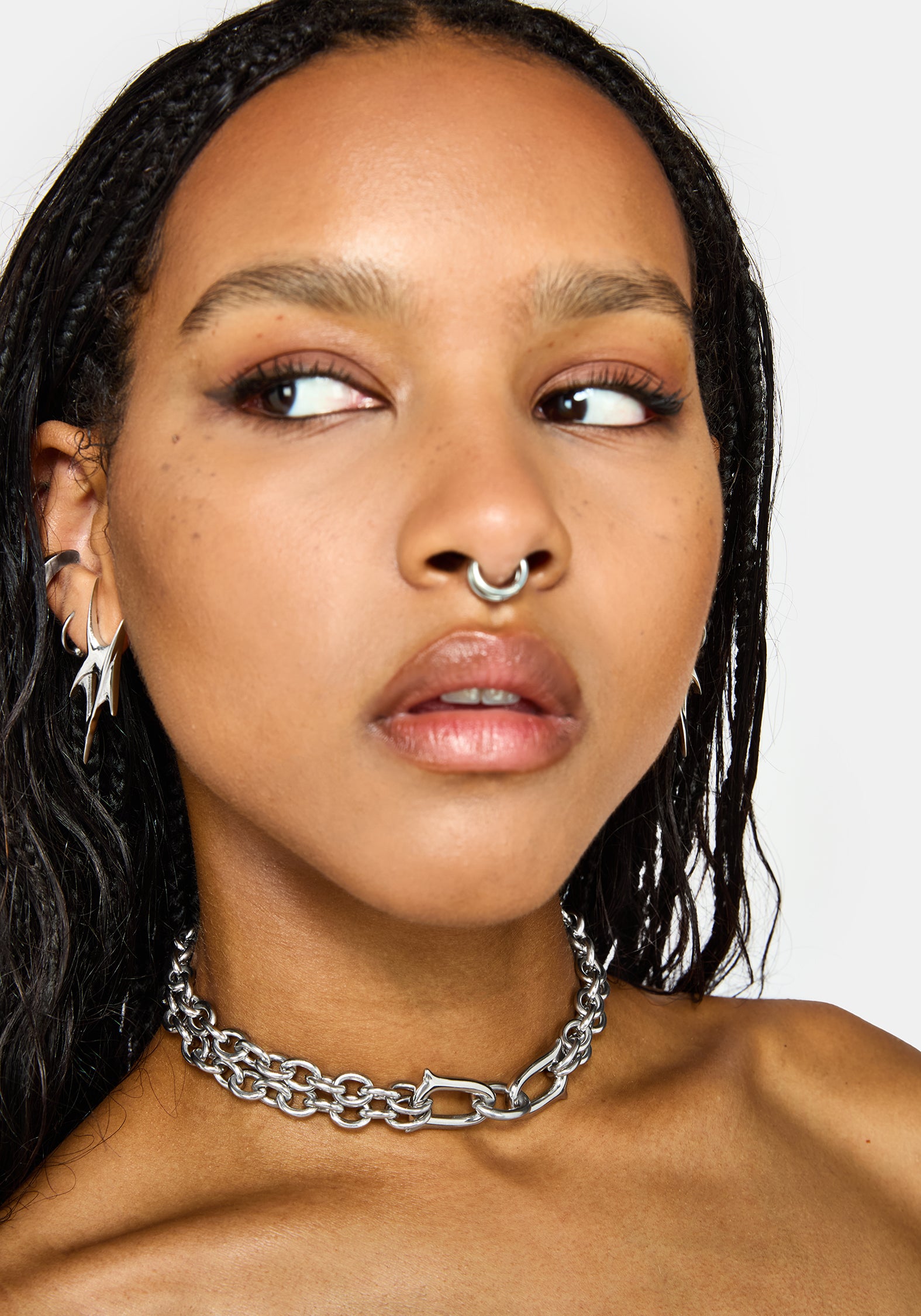 A close-up of a model wearing a chunky silver chain choker necklace with a lobster clasp, along with multiple silver piercings on her ears and nose.