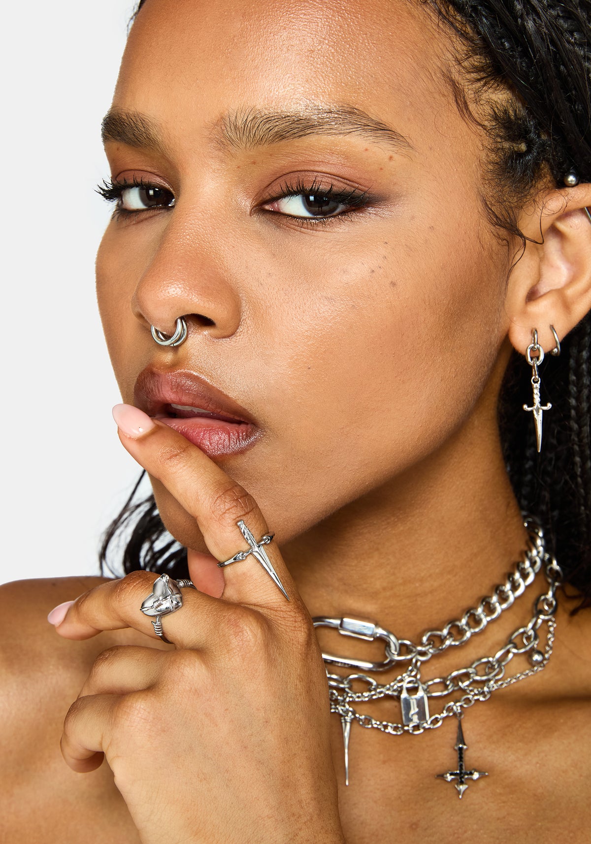 A close-up of a model wearing multiple silver-toned gothic jewelry pieces, including layered chain necklaces with a cross pendant, a ring, a nose ring, and a dangling earring, against a light gray background.