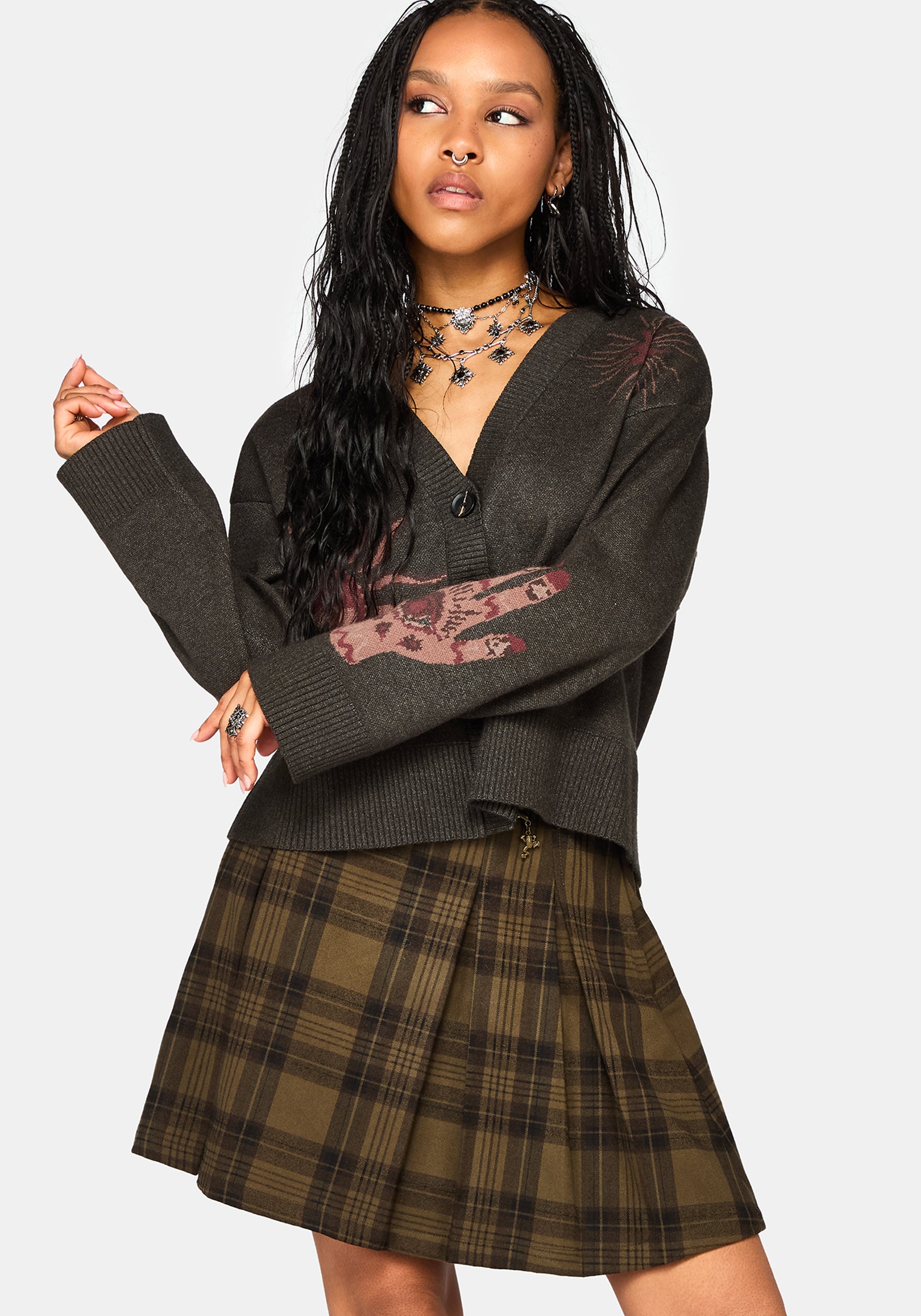 A woman modeling a dark brown knit cardigan with a graphic print on the sleeves and a brown plaid pleated mini skirt, set against a solid light gray background.