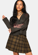 A woman modeling a dark brown knit cardigan with a graphic print on the sleeves and a brown plaid pleated mini skirt, set against a solid light gray background.